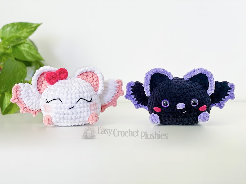Puede incluir: Dos juguetes de peluche de crochet, uno blanco con detalles rosas y un lazo rosa, y otro negro con detalles morados. Ambos juguetes tienen ojos bordados y una boca sonriente.