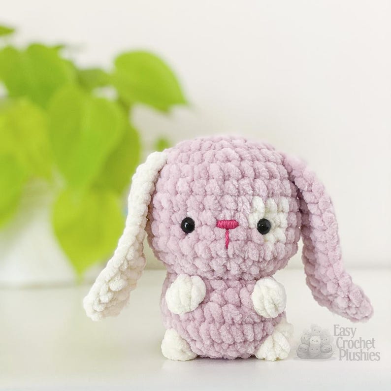 May include: A lavender crochet bunny plush toy with white detailing on the ears, paws, and face. It features black eyes and a pink embroidered nose. The text "Easy Crochet Plushies" is visible in the lower right corner.