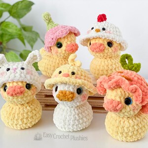 May include: A collection of five handmade crochet duck plushies, each with unique hats. The ducks are yellow with orange beaks and black eyes. Hats include a bunny, flower, chicken, and other whimsical designs. The plushies are displayed on a wooden surface.