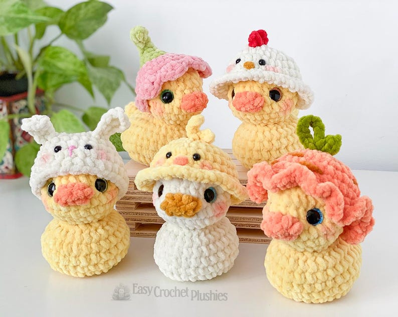 May include: A collection of five handmade crochet duck plushies, each with unique hats. The ducks are yellow with orange beaks and black eyes. Hats include a bunny, flower, chicken, and other whimsical designs. The plushies are displayed on a wooden surface.