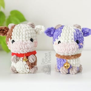 May include: Two handmade crochet cow plushies. One cow is brown, white, and red, with a gold bell. The other is purple, white, and brown, with a gold bell. Both have a cute, friendly design.