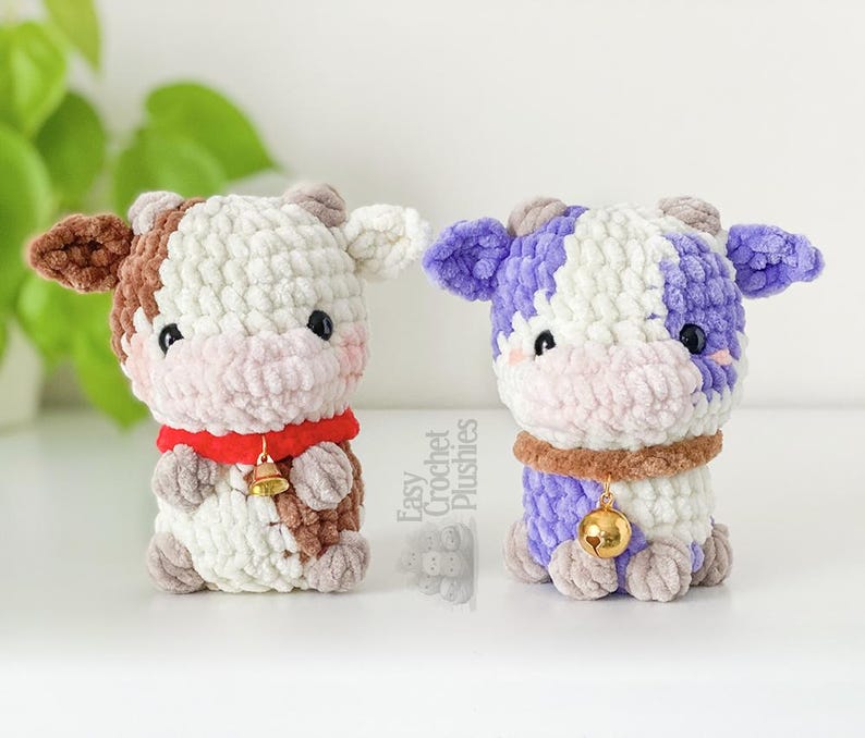 May include: Two handmade crochet cow plushies. One cow is brown, white, and red, with a gold bell. The other is purple, white, and brown, with a gold bell. Both have a cute, friendly design.