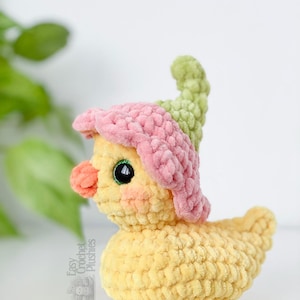 May include: A handmade yellow crochet duck plushie wearing a pink and green flower hat. The duck has a peach-colored beak and black eyes. The text "Easy Crochet Plushies" is visible on the side. The plushie is a whimsical and charming handmade item.