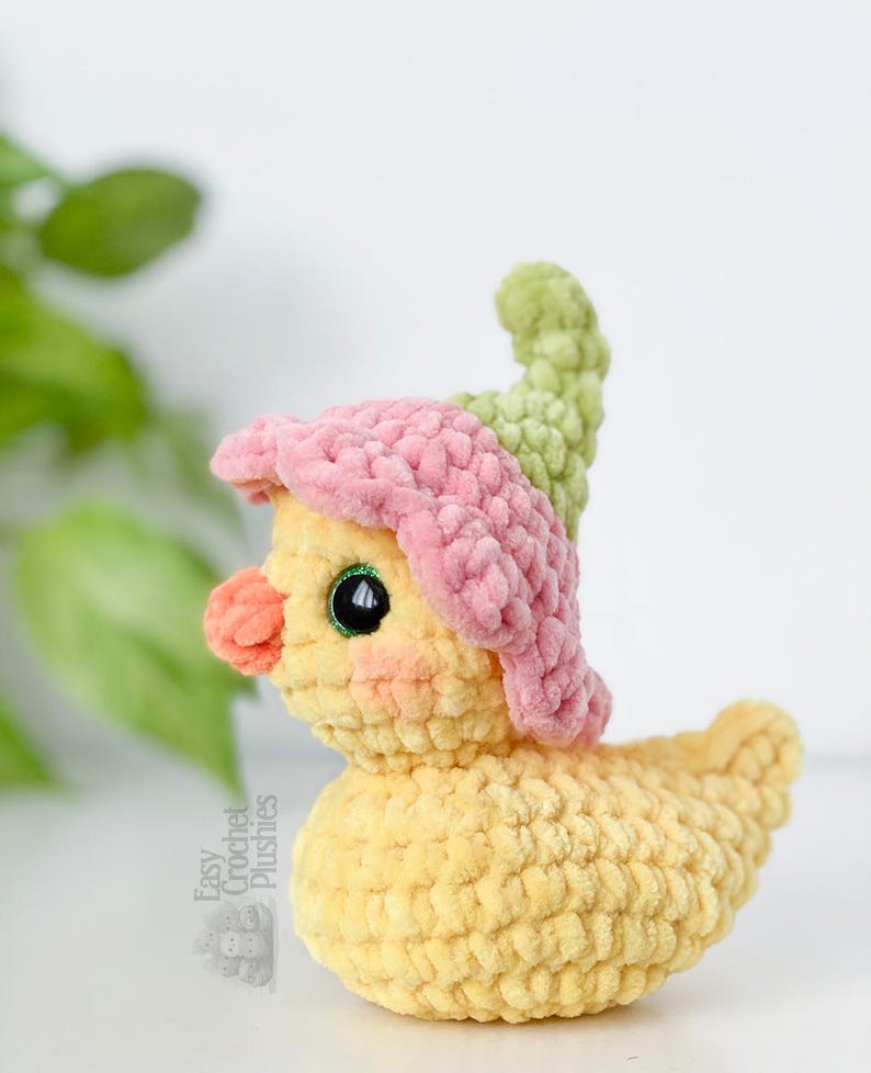 May include: A handmade yellow crochet duck plushie wearing a pink and green flower hat. The duck has a peach-colored beak and black eyes. The text "Easy Crochet Plushies" is visible on the side. The plushie is a whimsical and charming handmade item.