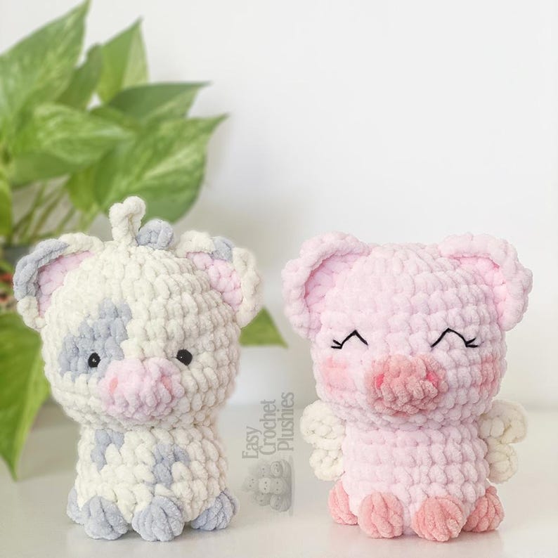 May include: Two handmade crochet pig plushies. One is white with grey spots and pink accents. The other is solid pink with rosy cheeks and a smiling expression. Both have small black eyes and are sitting upright.
