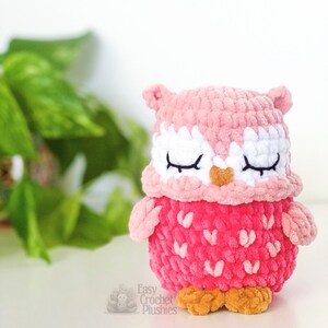 Puede incluir: Un juguete de peluche de b&uacute;ho de crochet rosa con los ojos cerrados y una nariz marr&oacute;n. El b&uacute;ho est&aacute; sentado sobre una superficie blanca con plantas verdes en el fondo.