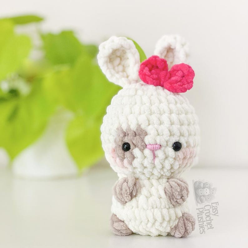May include: A handmade white crochet bunny plushie with a pink bow, grey accents, and black eyes. The bunny has a light pink nose and cheeks. The plushie is a soft, cuddly toy.