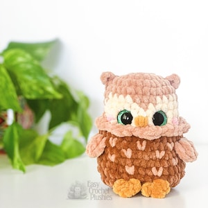 Puede incluir: Un juguete de peluche de b&uacute;ho de crochet marr&oacute;n y rosa con grandes ojos verdes. El b&uacute;ho tiene una barriga de crochet blanca y patas de crochet naranja. El juguete est&aacute; sentado sobre una superficie blanca.