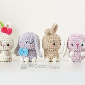 May include: Four handmade crochet bunny plushies in white, lavender, tan, and pink. Each bunny has a unique expression and features, including bows and contrasting ear and foot colours. The plushies are displayed on a white surface.