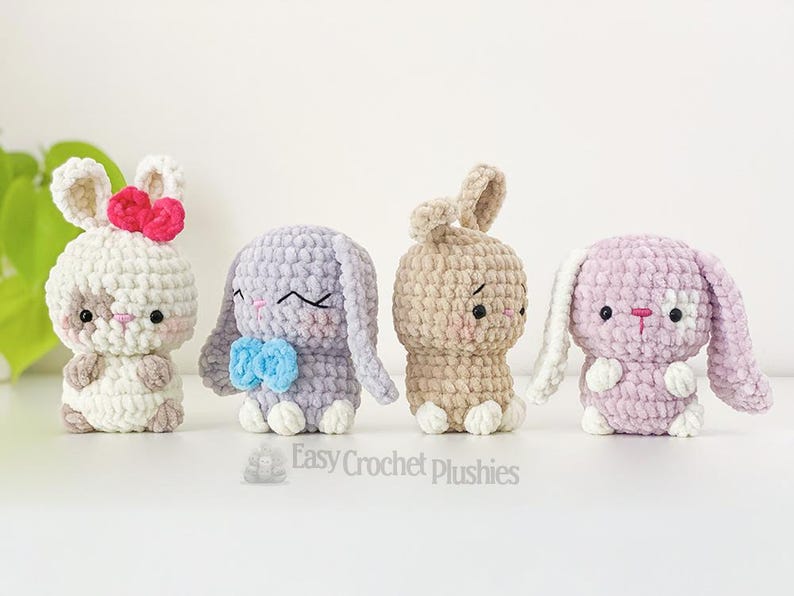 May include: Four handmade crochet bunny plushies in white, lavender, tan, and pink. Each bunny has a unique expression and features, including bows and contrasting ear and foot colours. The plushies are displayed on a white surface.