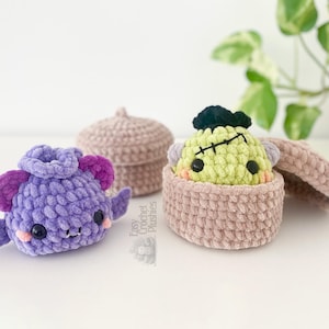May include: A purple crocheted bat plushie with dark purple ears and wings, positioned next to a light brown crocheted lidded container holding a light green crocheted monster plushie with black hair and a stitched scar on its forehead.