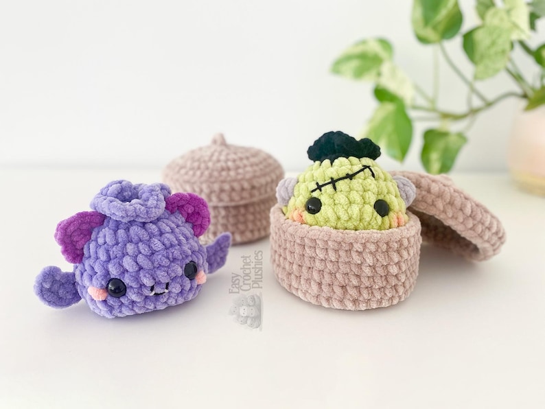 May include: A purple crocheted bat plushie with dark purple ears and wings, positioned next to a light brown crocheted lidded container holding a light green crocheted monster plushie with black hair and a stitched scar on its forehead.