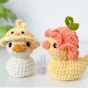 May include: Two handmade crochet duck plushies. One duck is white with a yellow hat and orange beak. The other duck is yellow with a pink head covering and a green stem. The plushies are on a white surface.