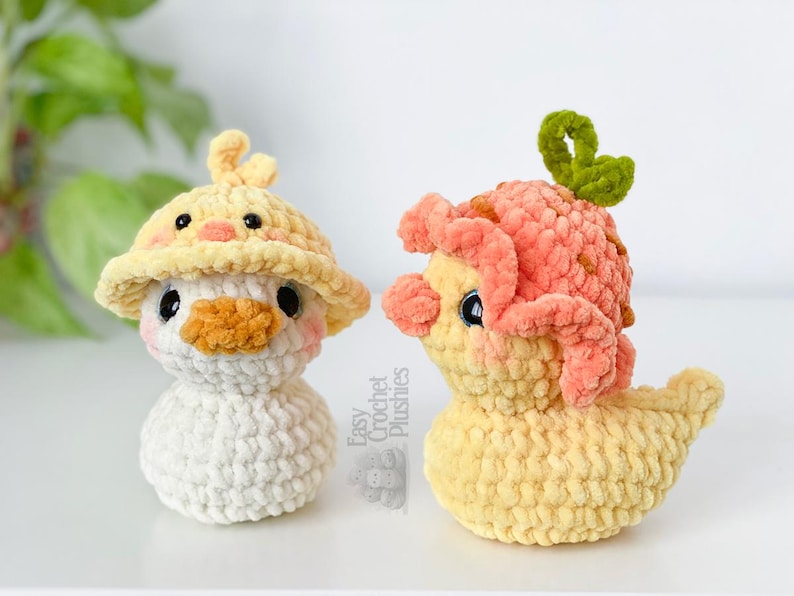 May include: Two handmade crochet duck plushies. One duck is white with a yellow hat and orange beak. The other duck is yellow with a pink head covering and a green stem. The plushies are on a white surface.
