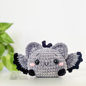 Puede incluir: Un juguete de peluche de murci&eacute;lago gris tejido a crochet con alas negras y mejillas rosadas. El juguete tiene grandes ojos negros y una peque&ntilde;a nariz negra. El juguete est&aacute; sentado sobre una superficie blanca.