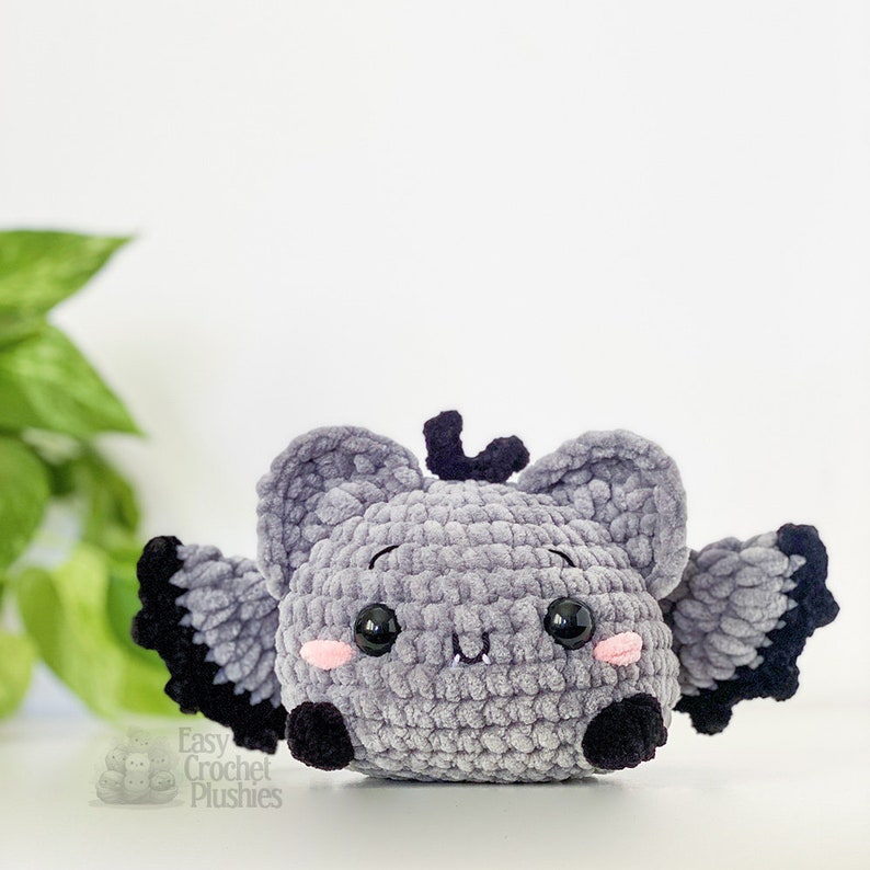 Puede incluir: Un juguete de peluche de murci&eacute;lago gris tejido a crochet con alas negras y mejillas rosadas. El juguete tiene grandes ojos negros y una peque&ntilde;a nariz negra. El juguete est&aacute; sentado sobre una superficie blanca.