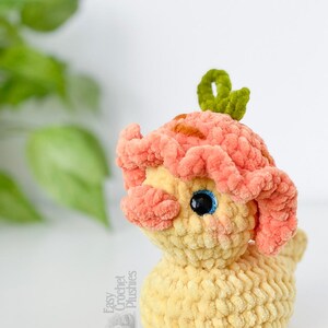 May include: A handmade crochet plushie in the shape of a whimsical creature. The plushie features a light yellow body, peach-colored head with brown accents, and a green leaf-like detail on top. The eyes are black with blue highlights. The text "Easy Crochet Plushies" is visible.