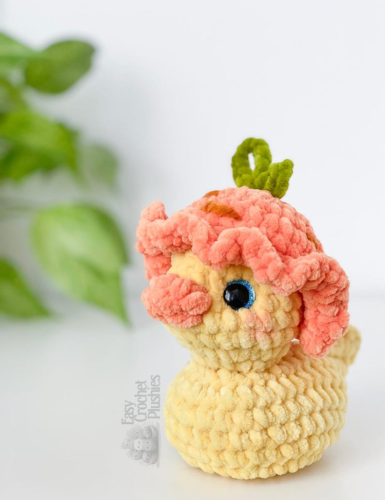 May include: A handmade crochet plushie in the shape of a whimsical creature. The plushie features a light yellow body, peach-colored head with brown accents, and a green leaf-like detail on top. The eyes are black with blue highlights. The text "Easy Crochet Plushies" is visible.