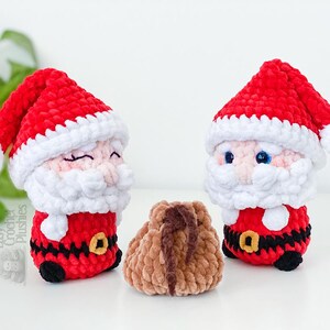 May include: Two crocheted Santa Claus plushies with red hats and white beards, and a small brown pouch. The Santa figures have black belts with gold buckles and black shoes. The plushies are on a white surface.