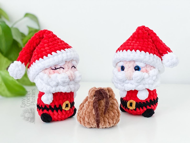 May include: Two crocheted Santa Claus plushies with red hats and white beards, and a small brown pouch. The Santa figures have black belts with gold buckles and black shoes. The plushies are on a white surface.