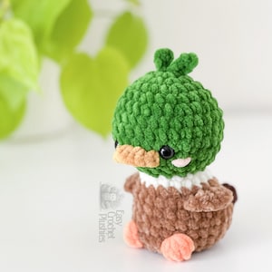 Peut inclure: Une petite peluche crochet&eacute;e &agrave; la main en forme de canard colvert. Elle pr&eacute;sente une t&ecirc;te ronde et textur&eacute;e verte avec des yeux de s&eacute;curit&eacute; noirs, un bec brun clair et des joues roses. Le corps est brun avec un col blanc et des pattes orange.