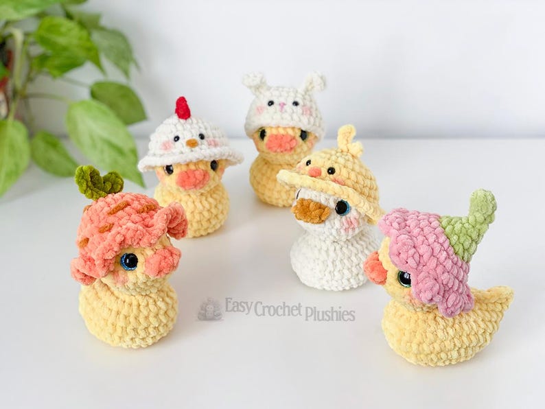 May include: Five handmade crochet duck plushies, each wearing a different hat. The ducks are yellow with orange beaks and blue eyes. Hats include a chicken, bunny, and mushroom design. The text "Easy Crochet plushies" is visible.