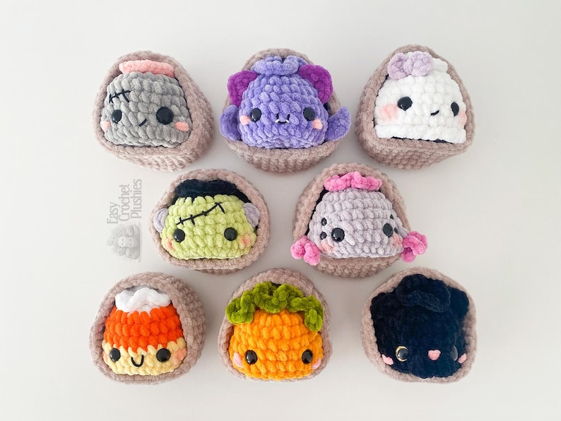 May include: Eight small, round, crocheted plushies in individual tan yarn baskets. The plushies feature various designs including a grey monster, purple creature, white ghost, green Frankenstein-like figure, grey spider, candy corn, pumpkin, and black cat.