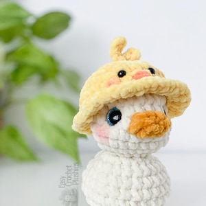 May include: A handmade crochet plushie of a duck wearing a yellow hat. The duck has a white body, an orange beak, and blue eyes. The hat is yellow with a duckling design. The plushie is soft and fluffy.
