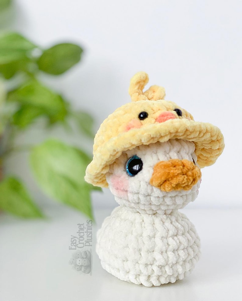 May include: A handmade crochet plushie of a duck wearing a yellow hat. The duck has a white body, an orange beak, and blue eyes. The hat is yellow with a duckling design. The plushie is soft and fluffy.