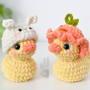 May include: Two handmade crochet plushies of yellow ducklings. One duckling wears a white bunny hat with pink accents, the other wears a peach-colored flower hat. Both have black eyes and peach-colored cheeks.