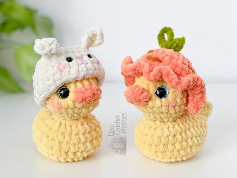 May include: Two handmade crochet plushies of yellow ducklings. One duckling wears a white bunny hat with pink accents, the other wears a peach-colored flower hat. Both have black eyes and peach-colored cheeks.