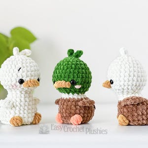 May include: Three handmade crochet duck plushies. Two are white with brown and tan accents, and one is green and brown. Each has black eyes and orange beaks. The plushies are displayed on a white surface.