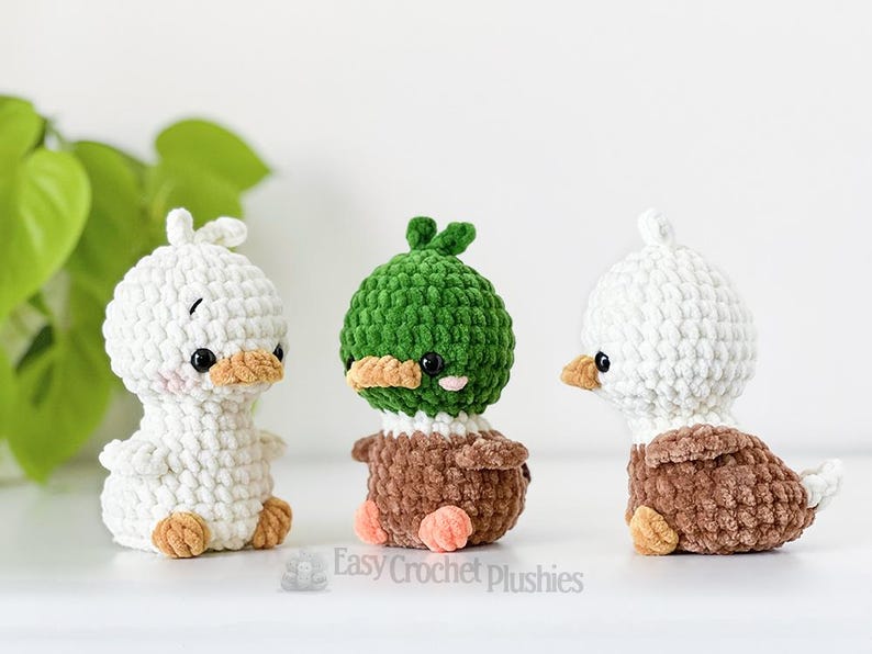 May include: Three handmade crochet duck plushies. Two are white with brown and tan accents, and one is green and brown. Each has black eyes and orange beaks. The plushies are displayed on a white surface.