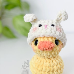 May include: A handmade yellow crochet duck wearing a white bunny hat with pink cheeks and a pink nose. The duck has black eyes and a textured, plush appearance. The text "Easy Crochet Plushies" is visible on the side.