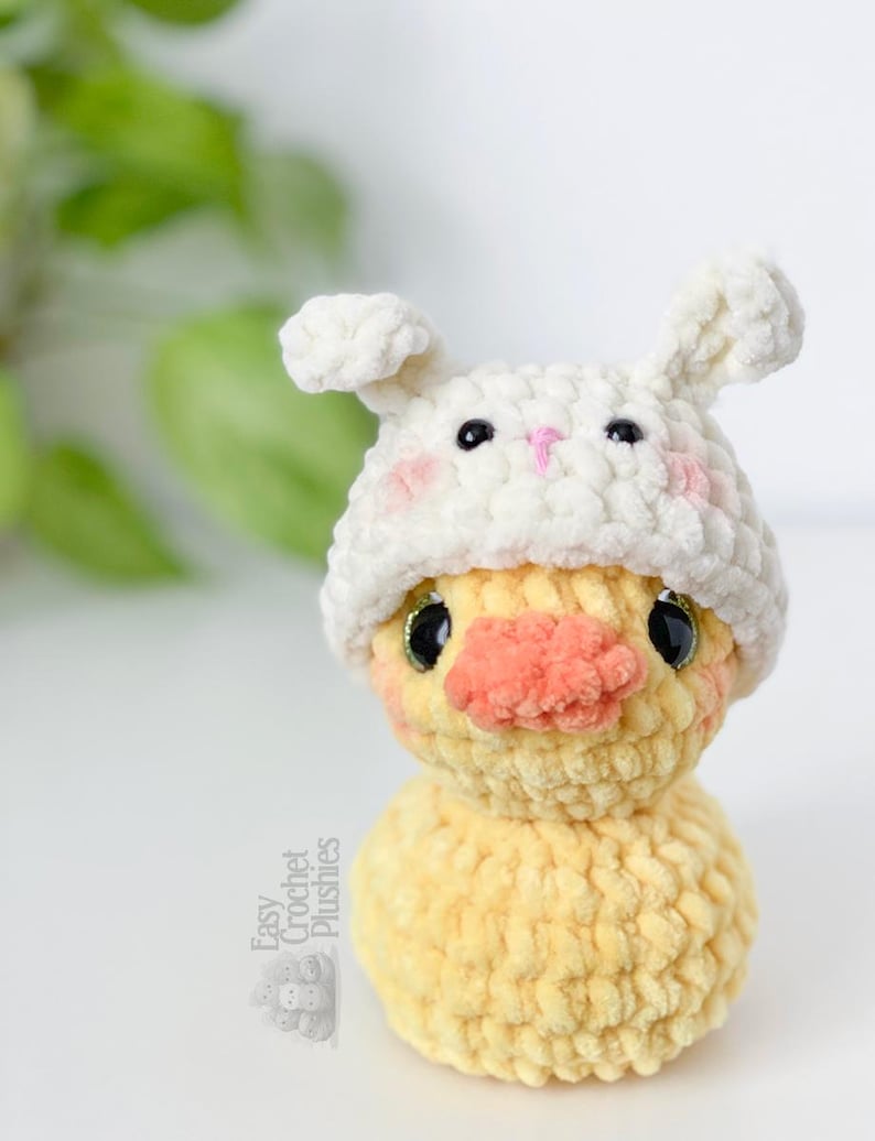 May include: A handmade yellow crochet duck wearing a white bunny hat with pink cheeks and a pink nose. The duck has black eyes and a textured, plush appearance. The text "Easy Crochet Plushies" is visible on the side.