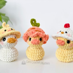 May include: Three handmade crochet duck plushies in yellow, white, and peach colors. Each duck wears a different hat: a yellow sun hat, a peach-colored mushroom cap, and a white hat with a red crest. The plushies have black eyes and orange beaks.