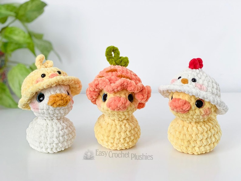 May include: Three handmade crochet duck plushies in yellow, white, and peach colors. Each duck wears a different hat: a yellow sun hat, a peach-colored mushroom cap, and a white hat with a red crest. The plushies have black eyes and orange beaks.