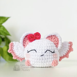 Puede incluir: Un juguete de peluche blanco tejido a crochet con orejas y alas rosas. El juguete tiene un lazo rosa en la cabeza y una cara linda y somnolienta con ojos negros y un peque&ntilde;o ce&ntilde;o fruncido. El juguete est&aacute; sentado sobre una superficie blanca.