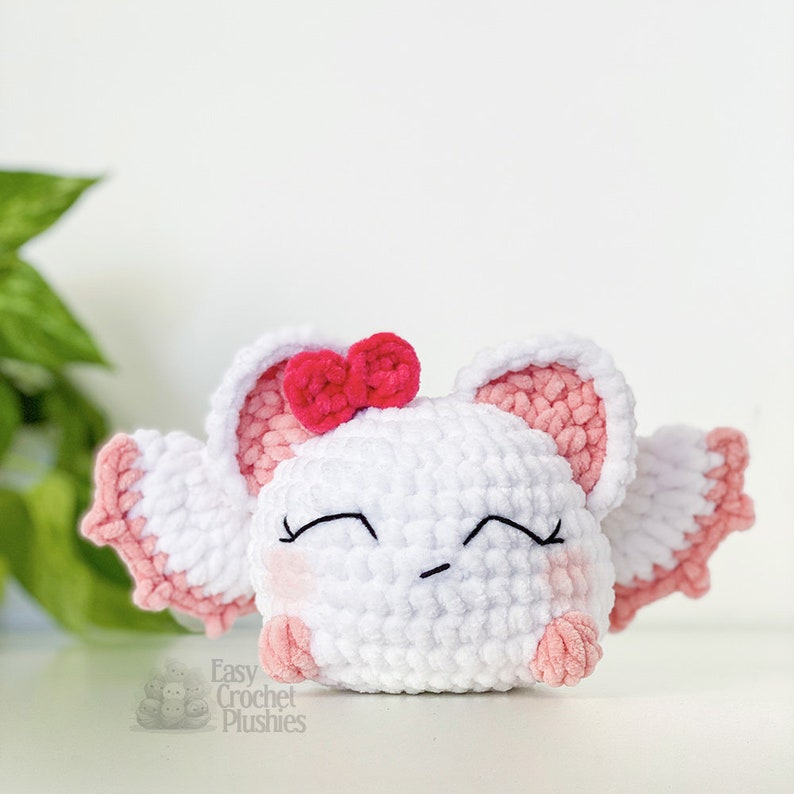Puede incluir: Un juguete de peluche blanco tejido a crochet con orejas y alas rosas. El juguete tiene un lazo rosa en la cabeza y una cara linda y somnolienta con ojos negros y un peque&ntilde;o ce&ntilde;o fruncido. El juguete est&aacute; sentado sobre una superficie blanca.