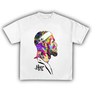 May include: White t-shirt featuring a colorful profile portrait. The portrait showcases a person with a white headband and beard, rendered in vibrant hues of blue, green, yellow, and purple. The word "KING" is written in black script.