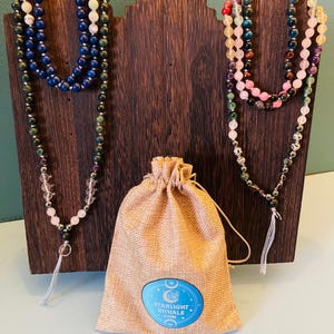 May include: Two beaded necklaces in shades of blue, green, and pink are displayed on a wooden stand. A tan drawstring bag with a blue and white logo is in front of the stand. The logo reads "Starlight Rituals".