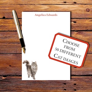 May include: A white notepad with the name "Angelica Edwards" printed in red. A silver and black fountain pen rests on the wooden surface. A sign reads "Choose from 50 Different Cat Images". A gray cat is pictured.