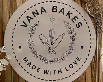 Op de afbeelding: Een rond beige bord met de zwarte tekst VANA BAKES bovenaan en MADE WITH LOVE onderaan. Het midden toont een zwarte lijntekening van een garde, spatel en deegroller binnen een bladerkrans met een klein hartje erboven.