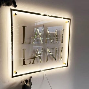 May include: Illuminated sign with the name "LAMI LANI" in gold lettering. The sign is rectangular with a gold border and gold corner accents. The sign is lit from behind with a soft, warm light.