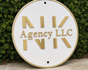 May include: A circular white sign with a gold border and gold lettering. The sign features the text "N I V" above "Agency LLC" and another "N I V" below, all in a metallic gold finish. There are two small circular holes at the top and bottom.