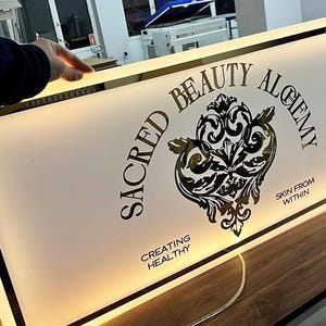 May include: Illuminated rectangular sign with the text "SACRED BEAUTY ALCHEMY" in black, along with a decorative emblem. The sign also includes the phrases "SKIN FROM WITHIN" and "CREATING HEALTHY". The sign has a gold border.