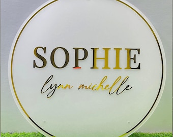 May include: A circular frosted acrylic sign featuring a gold border and gold lettering. The text reads SOPHIE in a bold serif font, with the names lynn michelle written below in a gold cursive script. The sign has small mounting holes.