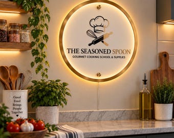May include: A circular wall-mounted sign with a gold frame and internal backlighting. The white center features a black graphic of a chef hat, whisk, and rolling pin, with the text THE SEASONED SPOON GOURMET COOKING SCHOOL & SUPPLIES.