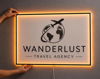 May include: A rectangular wall sign with a white face and black frame, featuring a black graphic of a globe with an airplane flying around it. The text reads WANDERLUST TRAVEL AGENCY in black capital letters. The sign emits a warm orange glow.