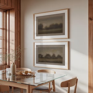 Könnte beinhalten: Zwei gerahmte Landschaftskunst-Drucke mit gedämpfter Farbpalette hängen an einer neutralen Wand. Das Kunstwerk zeigt eine verschwommene Darstellung von Bäumen und Feldern. Die Rahmen sind aus hellem Holz. Im Vordergrund befindet sich ein Esstisch mit Glasplatte und Holzstühlen.